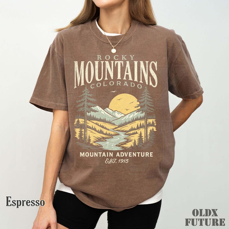 Puede incluir: Camiseta color espresso con un gr&aacute;fico de paisaje monta&ntilde;oso y el texto "Rocky Mountains Colorado Mountain Adventure Est. 1915". El dise&ntilde;o incluye un r&iacute;o, &aacute;rboles y un sol. La camiseta es de corte holgado.