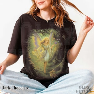 May include: Dark chocolate-colored t-shirt featuring a fantasy illustration of a fairy with butterfly wings, wearing a flowing white dress, and standing in a forest. A rabbit is at the fairy's feet. The text "Dark Chocolate" is at the bottom.
