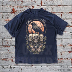 May include: Navy blue t-shirt featuring a graphic of a raven perched atop three skulls, with antlers and a butterfly. The design includes an orange moon and foliage. The text "OLDX FUTURE" is visible at the bottom.