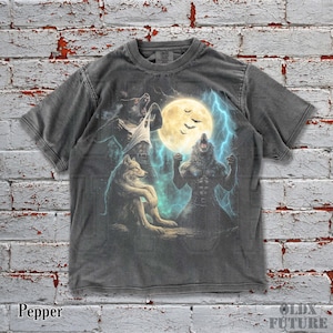 May include: Pepper-colored t-shirt featuring a graphic of wolves and a werewolf under a full moon with lightning. The design includes a seated wolf, a werewolf, and a wolf transforming into a werewolf. The shirt has a crew neck and short sleeves.
