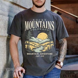 Puede incluir: Camiseta gris oscuro con un dise&ntilde;o de paisaje monta&ntilde;oso y el texto "Rocky Mountains Colorado Mountain Adventure Est. 1915". El gr&aacute;fico incluye un r&iacute;o, &aacute;rboles y un sol.