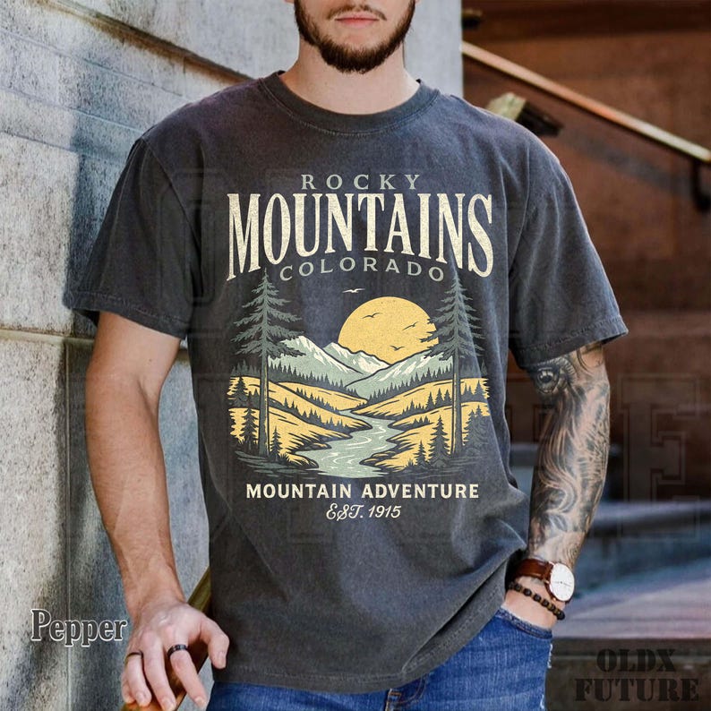 Puede incluir: Camiseta gris oscuro con un dise&ntilde;o de paisaje monta&ntilde;oso y el texto "Rocky Mountains Colorado Mountain Adventure Est. 1915". El gr&aacute;fico incluye un r&iacute;o, &aacute;rboles y un sol.