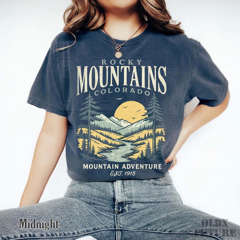 Puede incluir: Camiseta azul medianoche con un gr&aacute;fico de las Monta&ntilde;as Rocosas en Colorado. El dise&ntilde;o presenta una cordillera, un r&iacute;o, &aacute;rboles y un sol. El texto incluye "Rocky Mountains Colorado" y "Mountain Adventure Est. 1915".