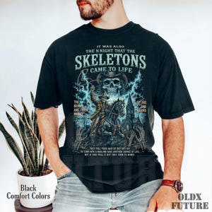 May include: A black t-shirt featuring a graphic print of a skeleton cowboy riding a horse in front of a church. The text reads: IT WAS ALSO THE N NIGHT THAT THE SKELETONS CAME TO LIFE, THE BONES ARE THEIR MONEY, SO ARE THE WORMS.