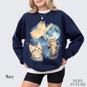 May include: Navy blue sweatshirt featuring a whimsical design of three kittens, a full moon, and a starry background. The sweatshirt has a crew neck and long sleeves. The word "Navy" is printed on the bottom left.