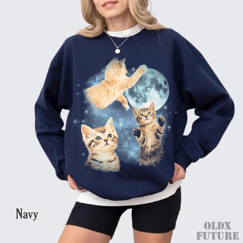 May include: Navy blue sweatshirt featuring a whimsical design of three kittens, a full moon, and a starry background. The sweatshirt has a crew neck and long sleeves. The word "Navy" is printed on the bottom left.