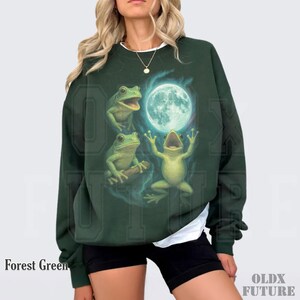 May include: Forest green sweatshirt featuring a whimsical illustration of three green frogs with a full moon in the background. The sweatshirt has the text "Forest Green" at the bottom left and "OLDX FUTURE" at the bottom right.