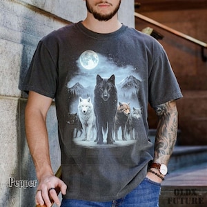 May include: Dark gray t-shirt featuring a wolf pack under a full moon. The graphic includes a black wolf, a white wolf, and other wolves in a mountain landscape. The shirt has a vintage, worn look.