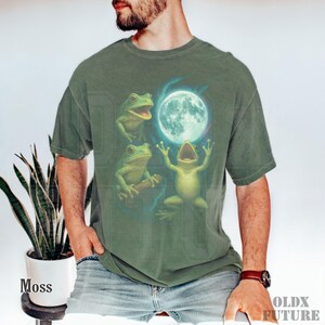 May include: Moss green t-shirt featuring a whimsical illustration of three green frogs with a full moon. One frog is holding a branch, and another is singing. The text "OLDX FUTURE" is printed on the lower right.