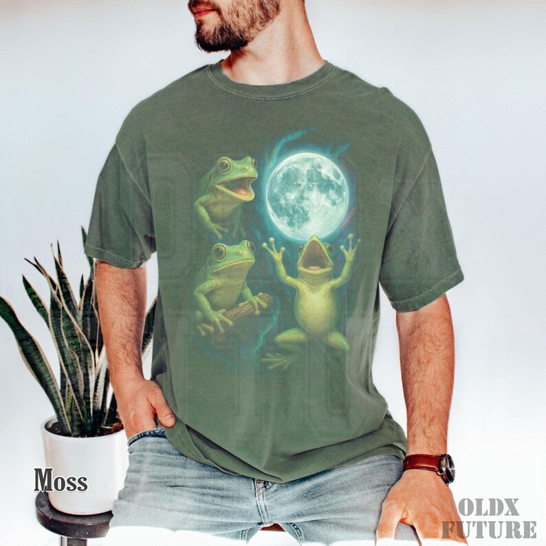May include: Moss green t-shirt featuring a whimsical illustration of three green frogs with a full moon. One frog is holding a branch, and another is singing. The text "OLDX FUTURE" is printed on the lower right.