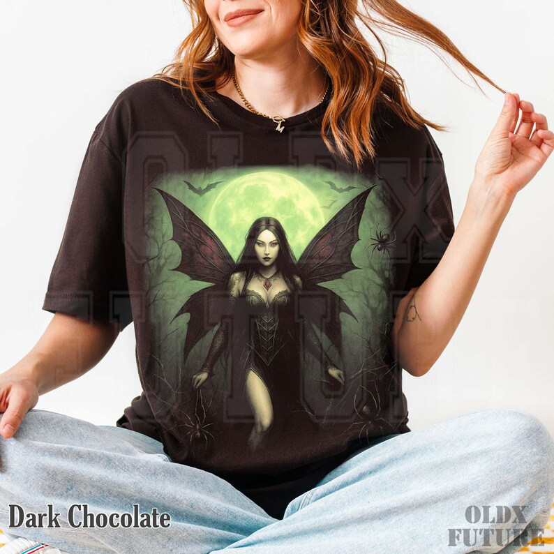 May include: Dark chocolate-colored t-shirt featuring a gothic fairy design. The graphic includes a woman with dark wings, a green moon, bats, and spiders. The text "Dark Chocolate" is at the bottom.