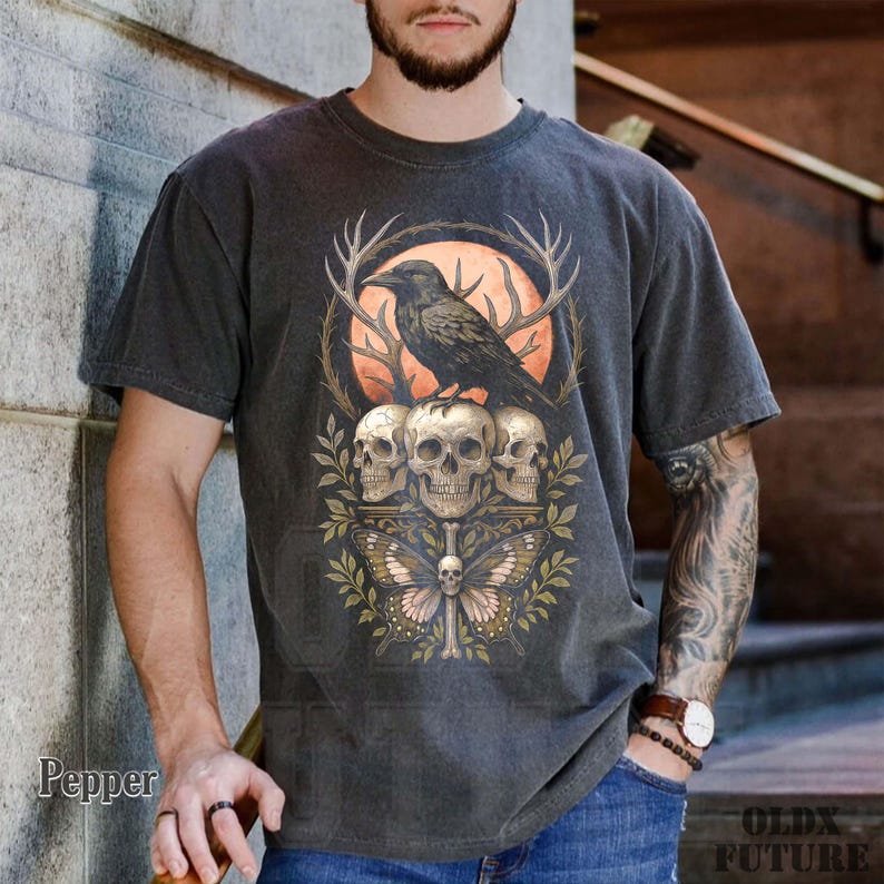 May include: Dark gray t-shirt featuring a detailed graphic. The design includes a raven perched above three skulls, a butterfly, and antlers, set against an orange backdrop. The shirt has a vintage, worn look.