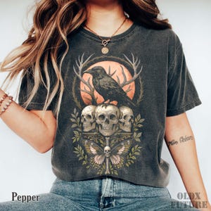 May include: Dark gray t-shirt featuring a detailed graphic of a raven perched above three skulls, a butterfly, and antlers. The design is set against a peach-colored circle. The word "Pepper" is printed at the bottom.