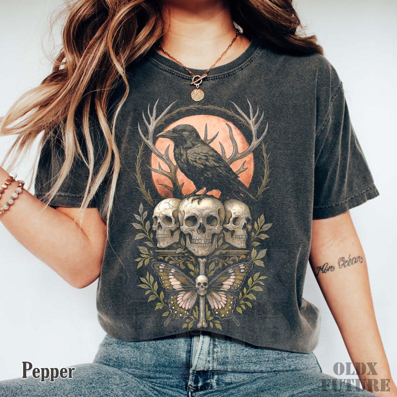 May include: Dark gray t-shirt featuring a detailed graphic of a raven perched above three skulls, a butterfly, and antlers. The design is set against a peach-colored circle. The word "Pepper" is printed at the bottom.