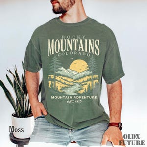 Puede incluir: Camiseta verde musgo con un dise&ntilde;o gr&aacute;fico de las Monta&ntilde;as Rocosas, Colorado, con un r&iacute;o, &aacute;rboles y un sol poniente. El texto dice "Rocky Mountains Colorado Mountain Adventure Est. 1915".