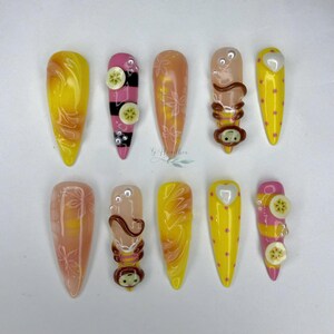 Könnte beinhalten: Ein Set aus zehn künstlichen Nägeln mit verschiedenen Designs. Die Nägel weisen Farben wie Gelb, Rosa und Pfirsich auf. Einige Nägel haben Bananenscheiben, Affenfiguren oder Blumenmuster. Die Nägel sind mandelförmig.
