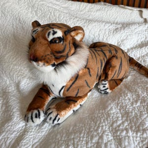 Puede incluir: Un tigre de peluche con pelaje naranja y marrón con rayas negras, pecho blanco y hocico blanco. El animal de peluche tiene ojos de color marrón oscuro, nariz rosa y bigotes, representado en posición tumbada con las patas delanteras extendidas.
