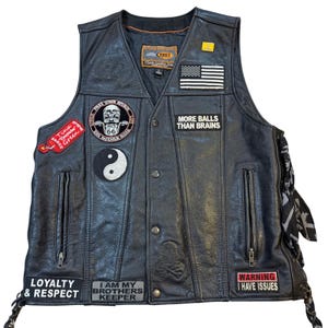 Könnte beinhalten: Schwarze Lederweste mit mehreren Aufnähern, darunter eine amerikanische Flagge, ein Route 66-Schädel und der Text „MORE BALLS THAN BRAINS“. Zusätzliche Aufnäher lauten „LOYALTY & RESPECT“ und „WARNING I HAVE ISSUES“.