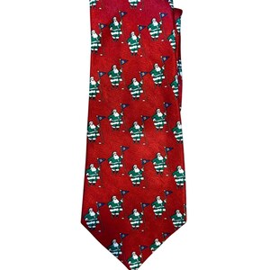 Op de afbeelding: Een rode stropdas met een herhaald patroon van kerstmanfiguren die vlaggen vasthouden. De kerstmanfiguren zijn groen en wit en de vlaggen zijn blauw. Een feestelijk accessoire.