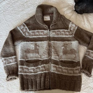 May include: A brown and beige knit zip-up cardigan featuring a shawl collar, long sleeves, and two front pockets. The garment displays a repeating pattern of deer and pine trees across the chest and sleeves, with a ribbed hem and cuffs.