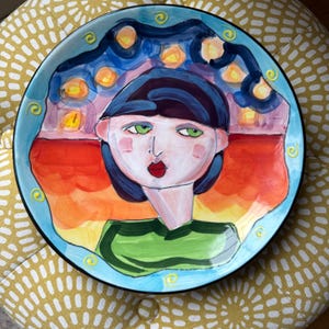 May include: A round ceramic plate featuring a hand-painted portrait of a person with dark blue hair, green eyes, and red lips. The figure wears a green shirt against a background of a sunset sky with orange, yellow, and blue swirling patterns.