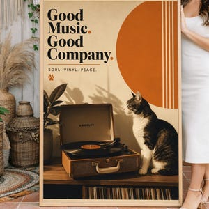Op de afbeelding: Een ingelijste poster met de tekst "Good Music. Good Company." heeft een retro-ontwerp met een oranje cirkel en verticale lijnen. Een vintage platenspeler en een zwart-witte kat staan voor de poster. De poster wordt door een persoon vastgehouden.