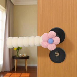 Könnte beinhalten: Ein Türgriff mit einem weißen Schaumstoffgriff und einer rosa blütenförmigen Abdeckung mit blauem Zentrum. Die Türbeschläge sind schwarz und die Tür ist aus Holz.