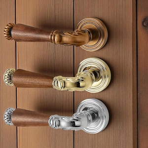 Może przedstawiać: Trzy ozdobne klamki do drzwi z drewnianymi uchwytami i dekoracyjnymi metalowymi akcentami w kolorze brązu, złota i srebra. Klamki są zamontowane na pionowych drewnianych drzwiach, prezentując ich design i wykończenie.