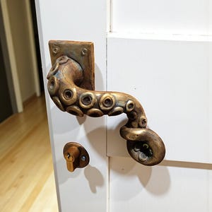 Puede incluir: Manija de puerta con forma de tentáculo de pulpo de color bronce y herrajes de cerradura a juego. La manija está montada sobre una placa cuadrada y presenta un diseño detallado de un tentáculo de pulpo. La cerradura también es de bronce.