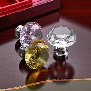May include: Three decorative cabinet knobs with faceted crystal tops in clear, yellow, and lavender. Each knob has a silver-colored metal base. The knobs are arranged on a dark wood surface, reflecting light and adding a touch of elegance.