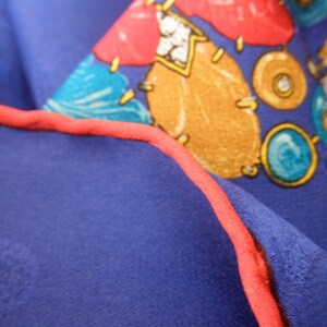 May include: Close-up of a blue fabric with a red trim. The fabric has a pattern of colorful shapes, including red, yellow, blue, and green. The shapes are abstract and resemble jewels or gemstones.