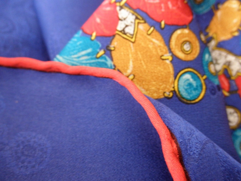May include: Close-up of a blue fabric with a red trim. The fabric has a pattern of colorful shapes, including red, yellow, blue, and green. The shapes are abstract and resemble jewels or gemstones.