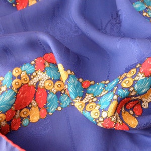 May include: A blue silk scarf with a colorful border. The border features red, yellow, blue, and white designs. The scarf is folded and draped over a surface.