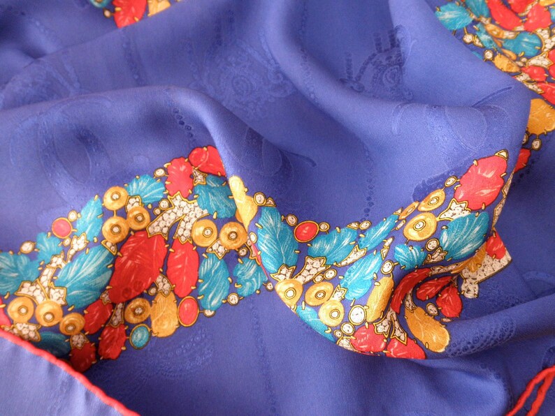 May include: A blue silk scarf with a colorful border. The border features red, yellow, blue, and white designs. The scarf is folded and draped over a surface.