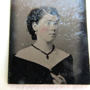 May include: A vintage black and white photograph of a woman. She wears a black dress with a low neckline and a black beaded necklace with a cross pendant. Her hair is styled in a bun, and she has a subtle blush on her cheeks.