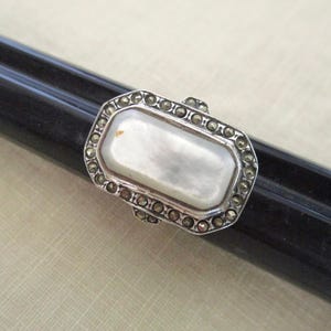 May include: A rectangular silver ring with a mother-of-pearl center stone. The ring is surrounded by small, round, silver-colored accents. The ring is displayed on a black cylindrical object against a light beige background. The ring is a piece of jewelry.