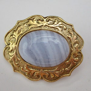 May include: A vintage brooch featuring an oval, light blue gemstone set in a gold-toned metal frame. The frame has an ornate, floral design. The brooch is a classic piece of jewelry.
