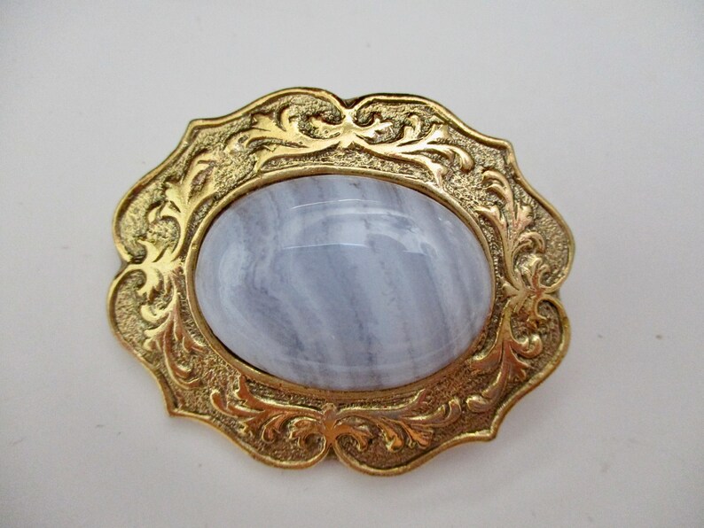 May include: A vintage brooch featuring an oval, light blue gemstone set in a gold-toned metal frame. The frame has an ornate, floral design. The brooch is a classic piece of jewelry.
