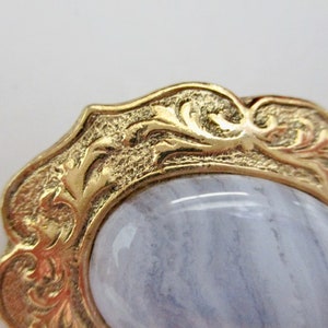 May include: Close-up of a gold-toned, ornate brooch with a light blue, oval-shaped stone. The gold frame features detailed, swirling patterns. The stone has a subtle, striped appearance.