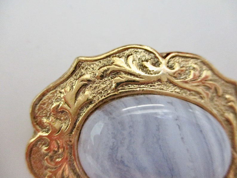 May include: Close-up of a gold-toned, ornate brooch with a light blue, oval-shaped stone. The gold frame features detailed, swirling patterns. The stone has a subtle, striped appearance.