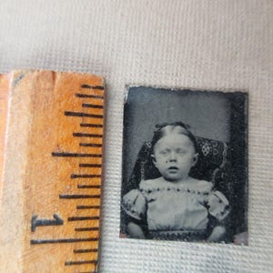 May include: A small, antique black and white photograph of a young child seated in a patterned chair. The child is wearing a dress with a ruffled neckline. A wooden ruler is positioned next to the photograph.