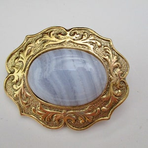 May include: A vintage brooch with a gold-tone filigree frame. The frame surrounds a large, oval, pale blue gemstone. The brooch has an ornate, decorative design, and is set against a white background.