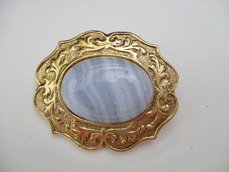 May include: A vintage brooch with a gold-tone filigree frame. The frame surrounds a large, oval, pale blue gemstone. The brooch has an ornate, decorative design, and is set against a white background.