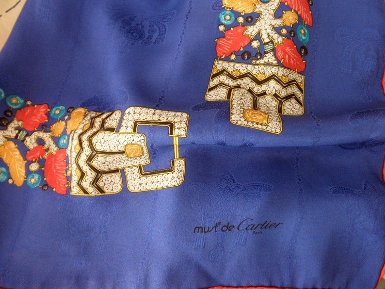 May include: A blue silk scarf with a white, black, gold, and red design. The design features a repeating pattern of a jeweled buckle. The scarf has a red border and the words 'must de Cartier Paris' in black text.