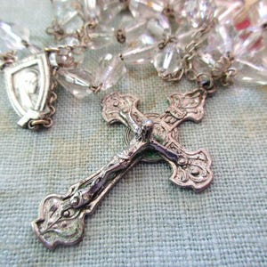 May include: A silver-toned rosary with clear, faceted beads and a detailed crucifix. The crucifix features an ornate design with the figure of Jesus Christ. A small shield-shaped pendant with a religious figure is also visible.