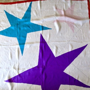 May include: White square scarf with a red border, featuring large, colorful star designs. The stars are in shades of blue, purple, and a faded pink, creating a bold and eye-catching pattern. The scarf is likely made of a lightweight fabric.