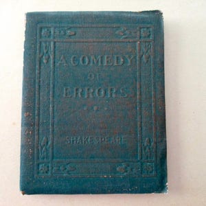 May include: A vintage book titled "A Comedy of Errors" by Shakespeare. The cover is a weathered teal color with embossed lettering and decorative borders. The book appears to be an antique.