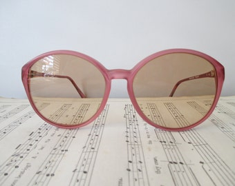 May include: Pink-framed sunglasses with brown-tinted lenses. The glasses have a rounded shape and thin, brown arms. The text "BOSTON EYE" is visible on the right arm. The sunglasses are resting on a sheet of vintage sheet music.
