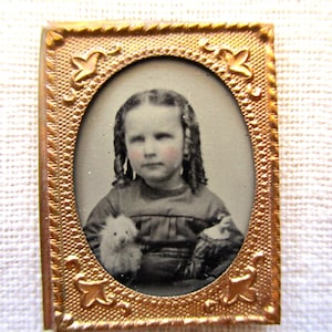 May include: Antique oval portrait of a young person in a gold-colored frame. The portrait is in black and white, with the subject holding a small stuffed animal. The frame has decorative floral accents.