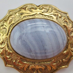 May include: A vintage gold-toned brooch with an oval, pale blue gemstone center. The gemstone has a marbled appearance. The brooch has an ornate, floral design around the gemstone.